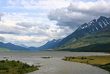 A large body of water, much longer than wide, lies at the base of mountains with vestiges of snow in their higher declivities. Vegetation is sparse. The mountains rise to meet a sky filled mostly with puffy white or gray clouds.