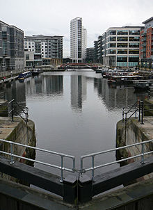 A night-time scene of a dock containing a number of moored canal-boats to left and right and railings around the edges. At the front is part of a lock gate and steps leading down to the water. Around most of the dock are multi-storey modern buildings, some with lighted ground-floors and seats and decorative objects outside. The most prominent of these, at the far end, is a twenty-storey building with curving fa&ccedil;ades.